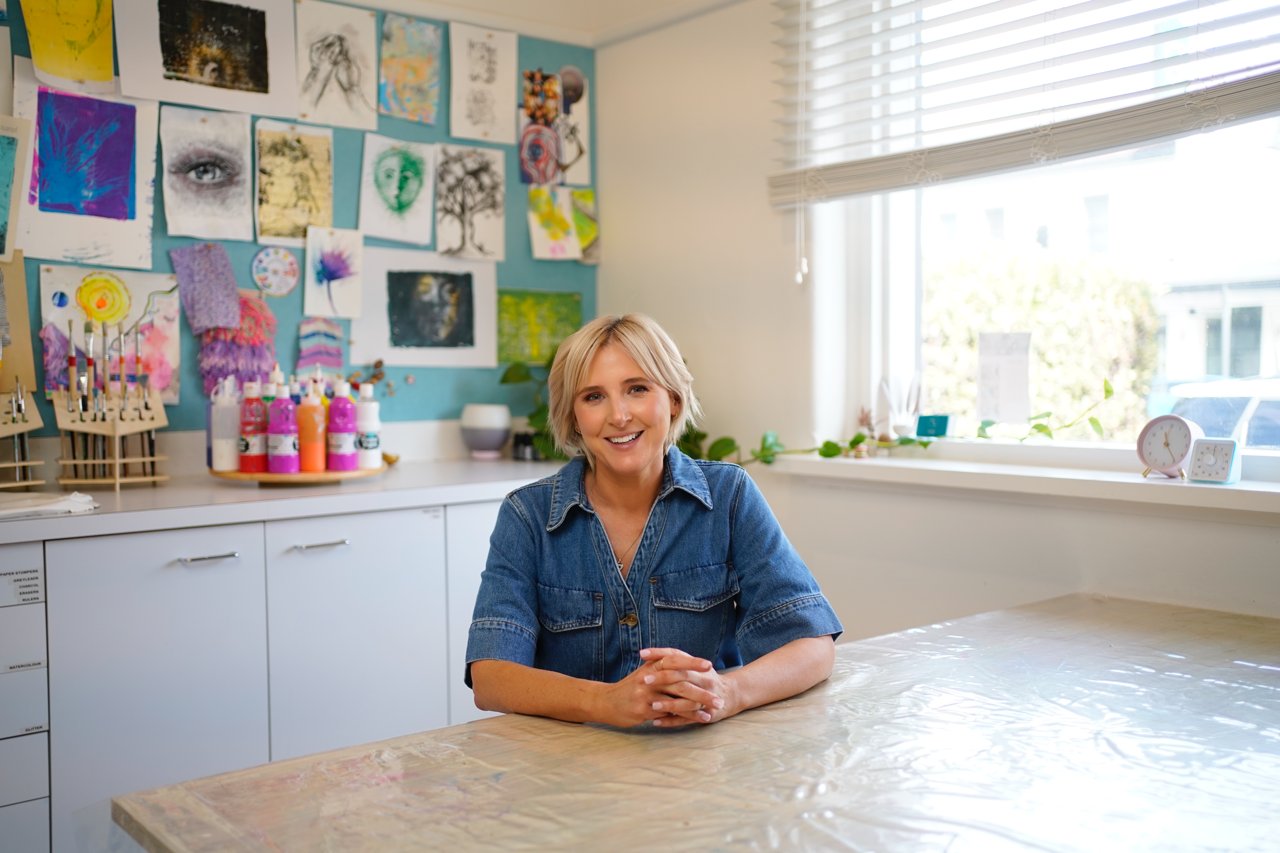 Orly Appel seated at the studio art table, with the client artwork wall behind her